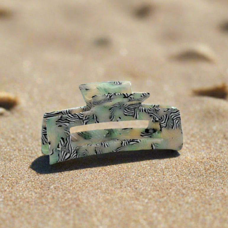 Zebra Haze rectangular hair claw clip made from plant-based cellulose acetate, side view on beach sand. Translucent sage green base with black-and-white zebra swirl pattern.