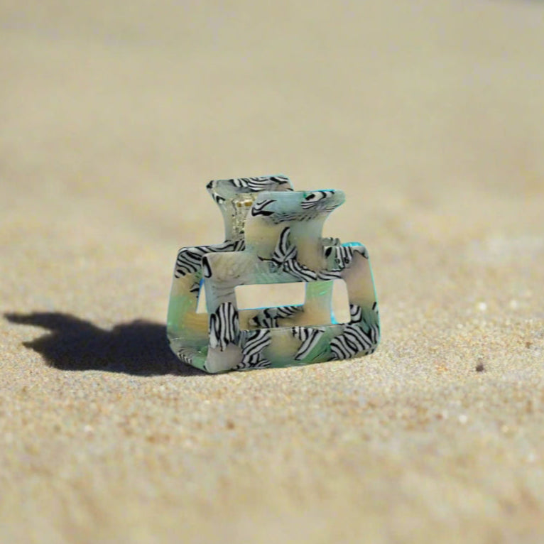 Zebra Haze rectangular hair claw clip made from plant-based cellulose acetate, side view on beach sand. Translucent sage green base with black-and-white zebra swirl pattern.