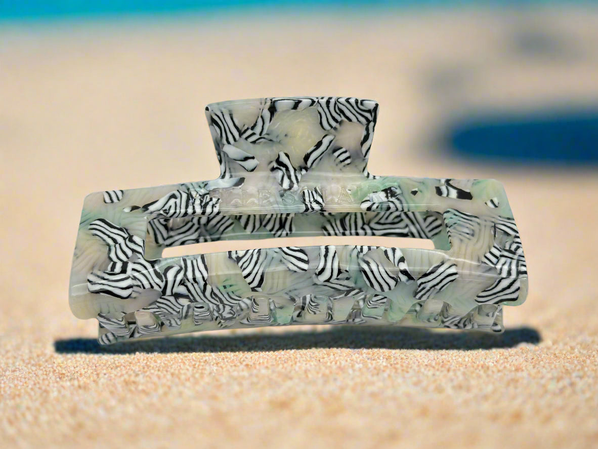Close-up of Zebra Haze hair clip showing claw teeth, resting on beach sand. Unique patterned acetate with a zebra-inspired design and semi-transparent green-beige tones.