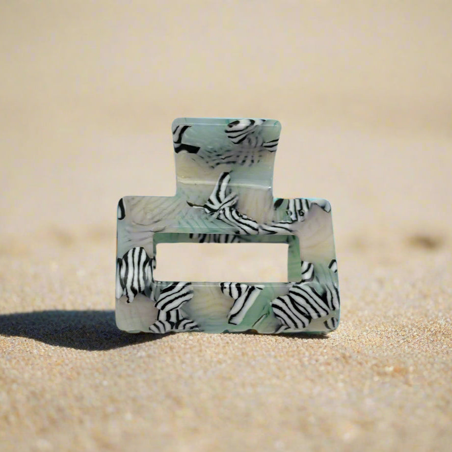 Zebra Haze claw clip in front view, lying on beach sand. Sleek rectangular shape with bold black-and-white zebra stripes over a soft sage-toned base.