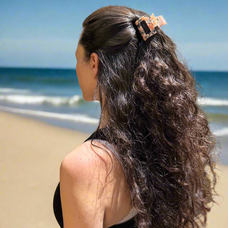 Woman with long brown hair styled in a half-up look wearing the Peach Gloss hair claw clip, standing at the beach in natural light.