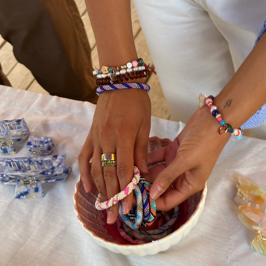 A close-up of a person's hands wearing and displaying colorful braided bracelets in a small light pink bowl, with hair clips arranged on a white tablecloth nearby.
