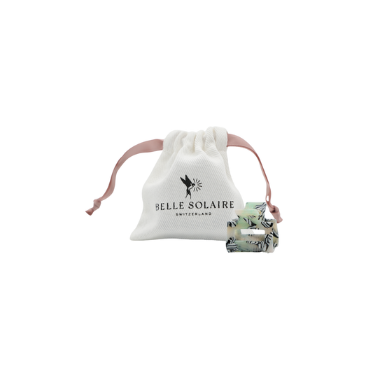 A light green and black and white stripped hair claw clip displayed next to its white drawstring pouch, which features the black printed logo 'BELLE SOLAIRE SWITZERLAND'.