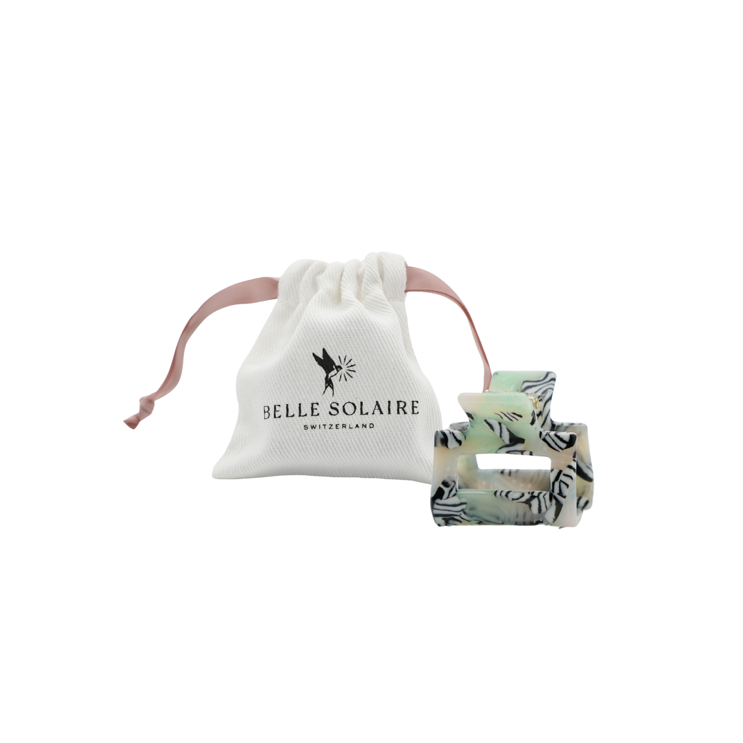 A light green and black and white striped hair claw clip displayed next to its white drawstring pouch, which features the black printed logo 'BELLE SOLAIRE SWITZERLAND'.