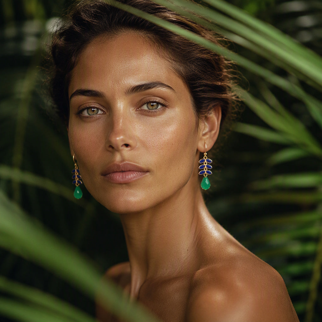 A close-up portrait of a woman with green teardrop earrings, framed by lush green palm leaves against a dark background.
