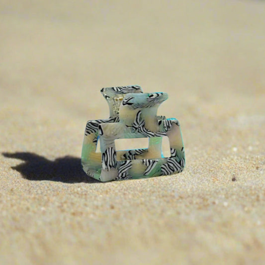Zebra Haze rectangular hair claw clip made from plant-based cellulose acetate, side view on beach sand. Translucent sage green base with black-and-white zebra swirl pattern.