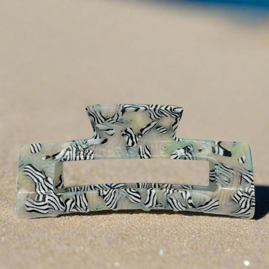 Zebra Haze claw clip in front view, lying on beach sand. Sleek rectangular shape with bold black-and-white zebra stripes over a soft sage-toned base.