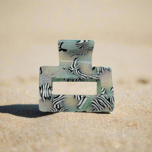 Zebra Haze claw clip in front view, lying on beach sand. Sleek rectangular shape with bold black-and-white zebra stripes over a soft sage-toned base.