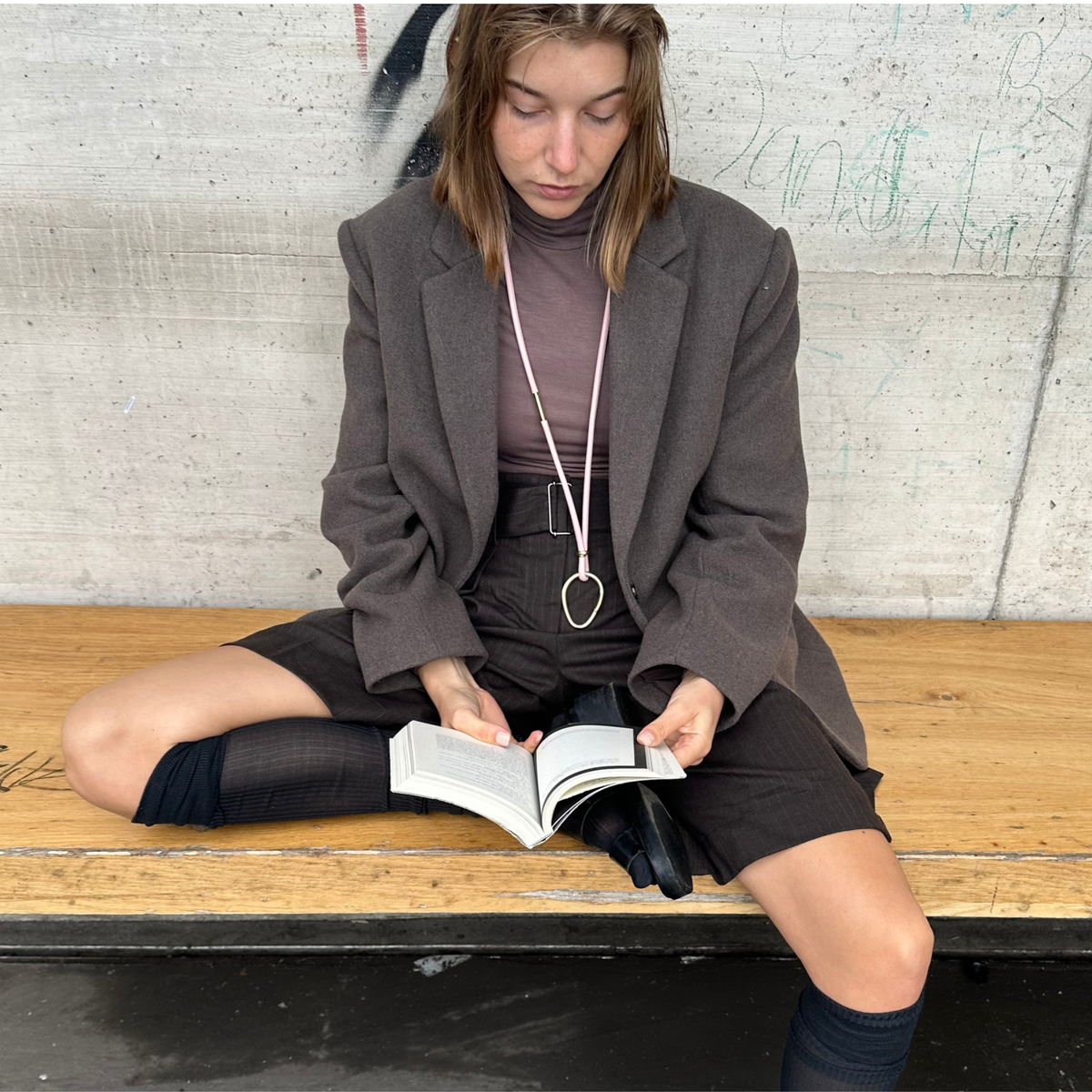 Woman reading a book on a bench with a textured wall background wearing a multipurpose leather strap with a large messing ring attached.
