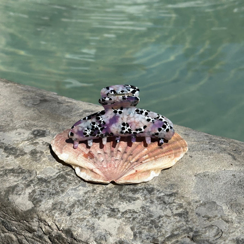 Hair claw clip with a  purple and pulkadot pattern on a shell lying on a rock by a body of water